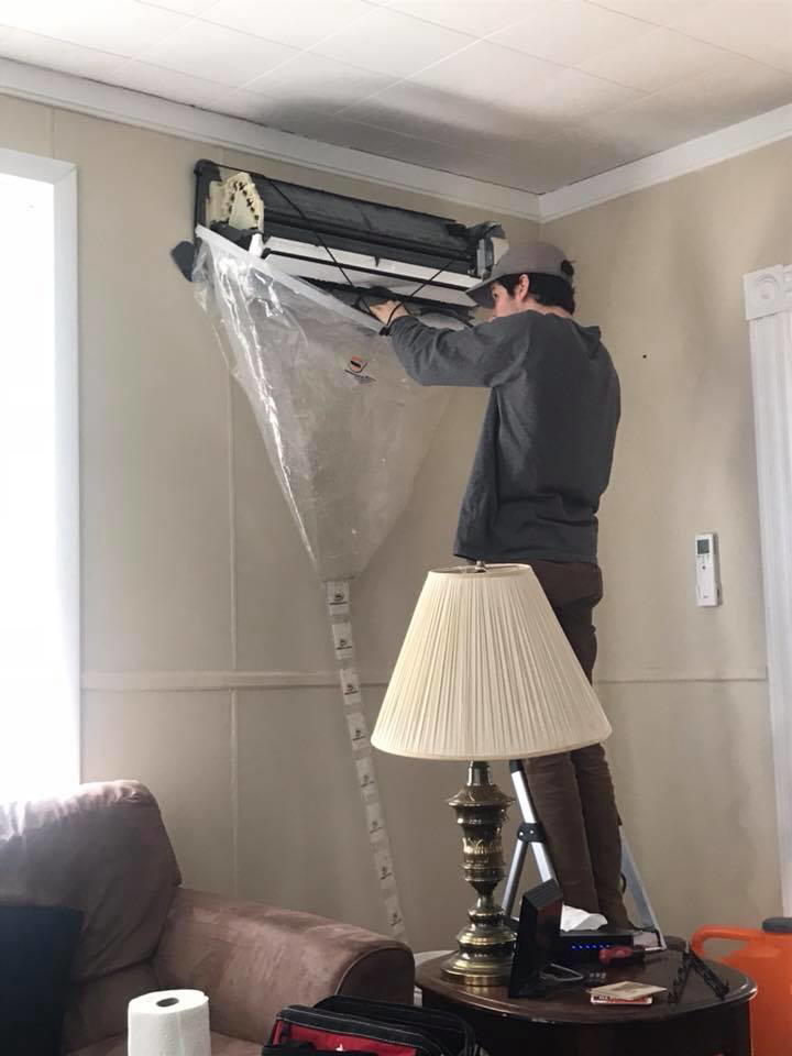 A technician on a ladder cleaning a mini-split heat pump unit with a protective cover at a client's home by Winks Heat Pump Cleanings in Bangor, ME