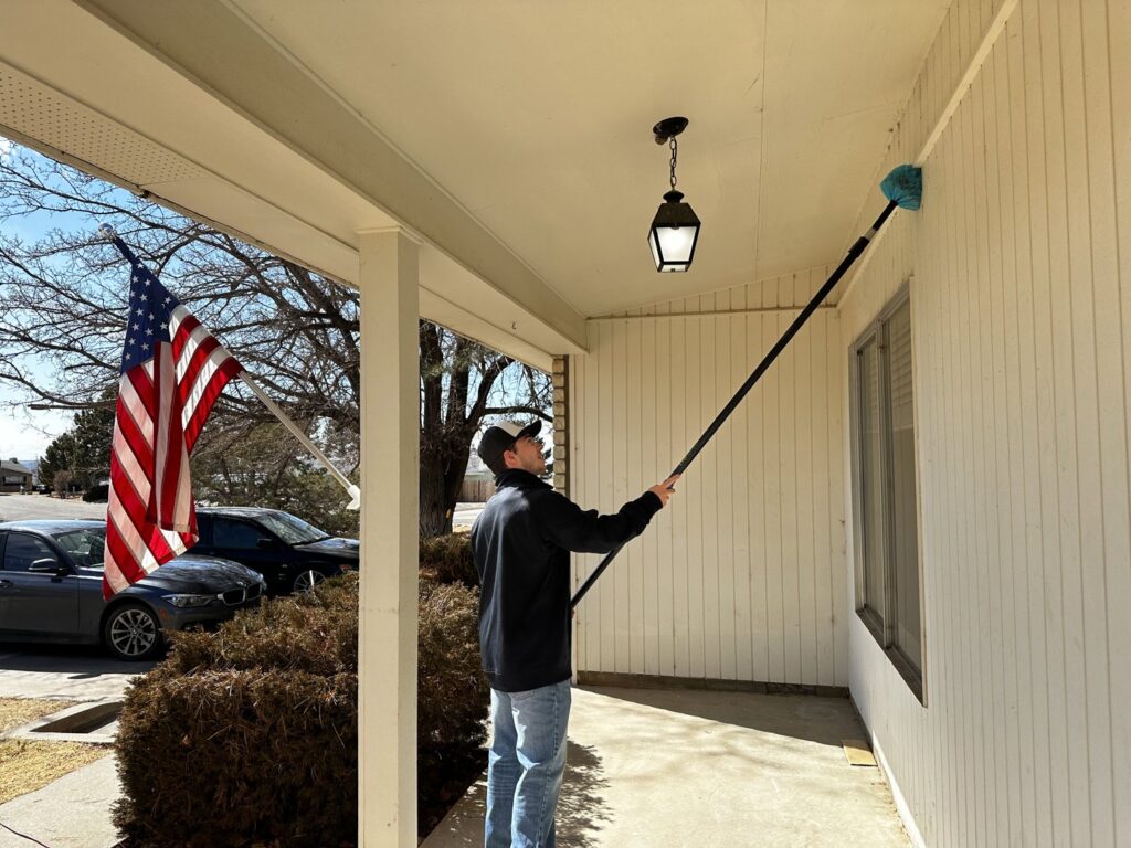 A Box Pest Control technician cleaning cobwebs from a porch ceiling with a long brush in Farmington, NM.