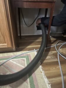 A technician from Boston Air Ducts Cleaning using a vacuum hose to clean a vent located under a kitchen cabinet in Newton, MA.