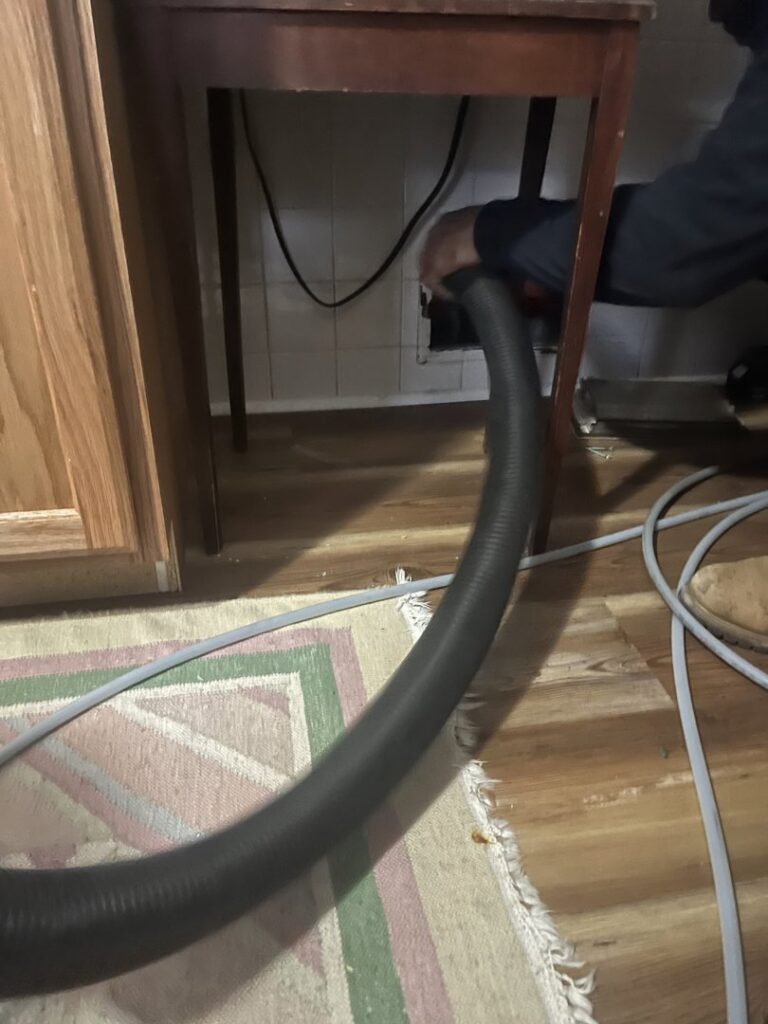A technician from Boston Air Ducts Cleaning using a vacuum hose to clean a vent located under a kitchen cabinet in Newton, MA.