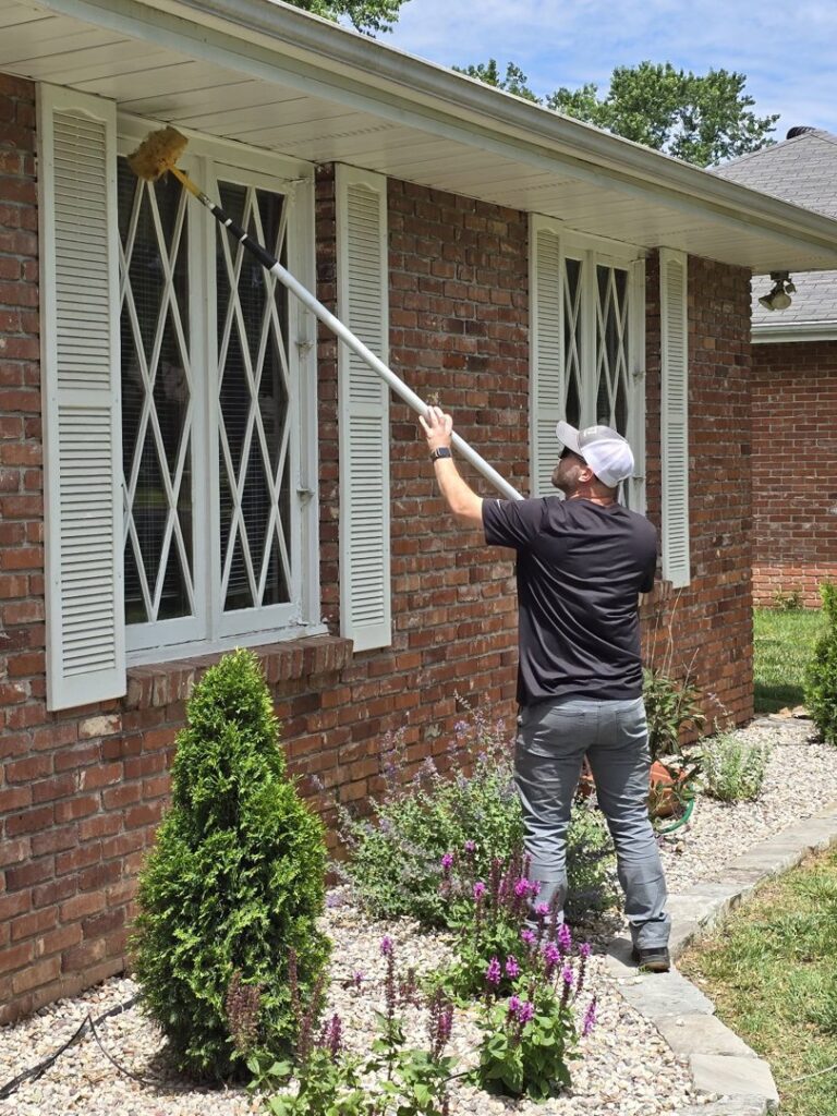 A pest control technician from Titan Pest Pro - Springfield using a long brush to clean around windows of a brick house in Springfield, MO.