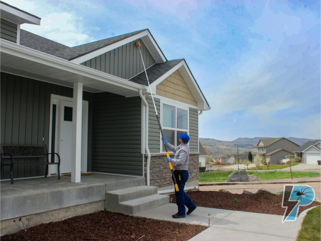 A Prompt Pest Solutions technician clearing spider webs from house eaves with a long pole in Pocatello, ID.