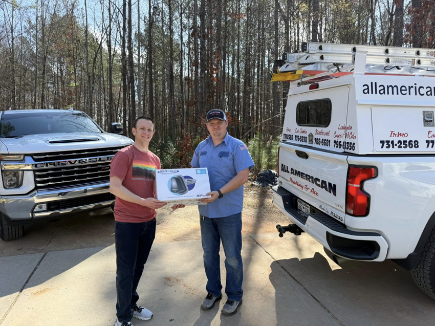 A technician and customer holding an HVAC product next to a company truck for All American Heating and Air in Warner Robins, GA.
