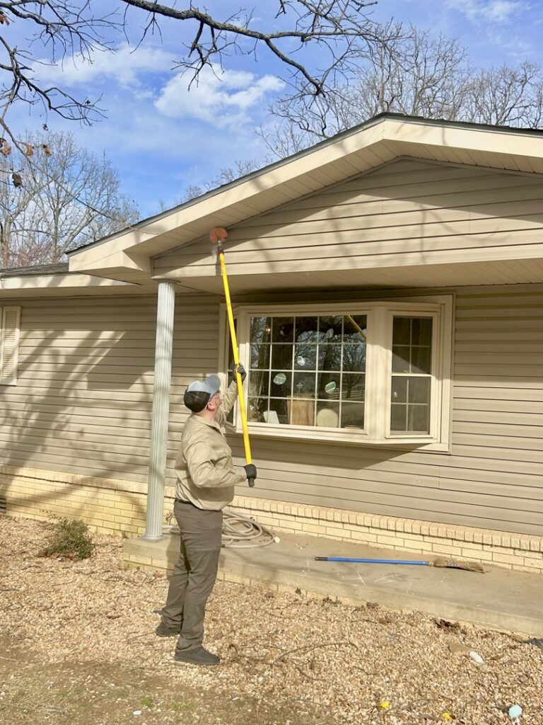 A Diamond Pest Solutions technician in Conway, AR, performing pest control by de-webbing the eaves of a house with a long pole.