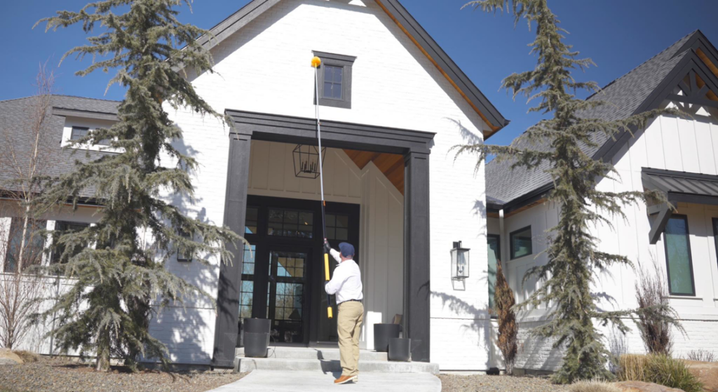 A Hemi Pest Control technician using a long pole to dust or remove webs from the eaves of a house in Meridian, ID.