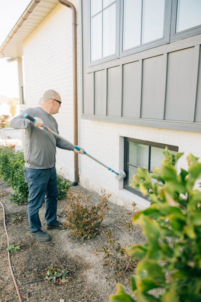 A technician performing exterior pest treatment on a house wall and window for Thrive Pest Management in Sandy, UT