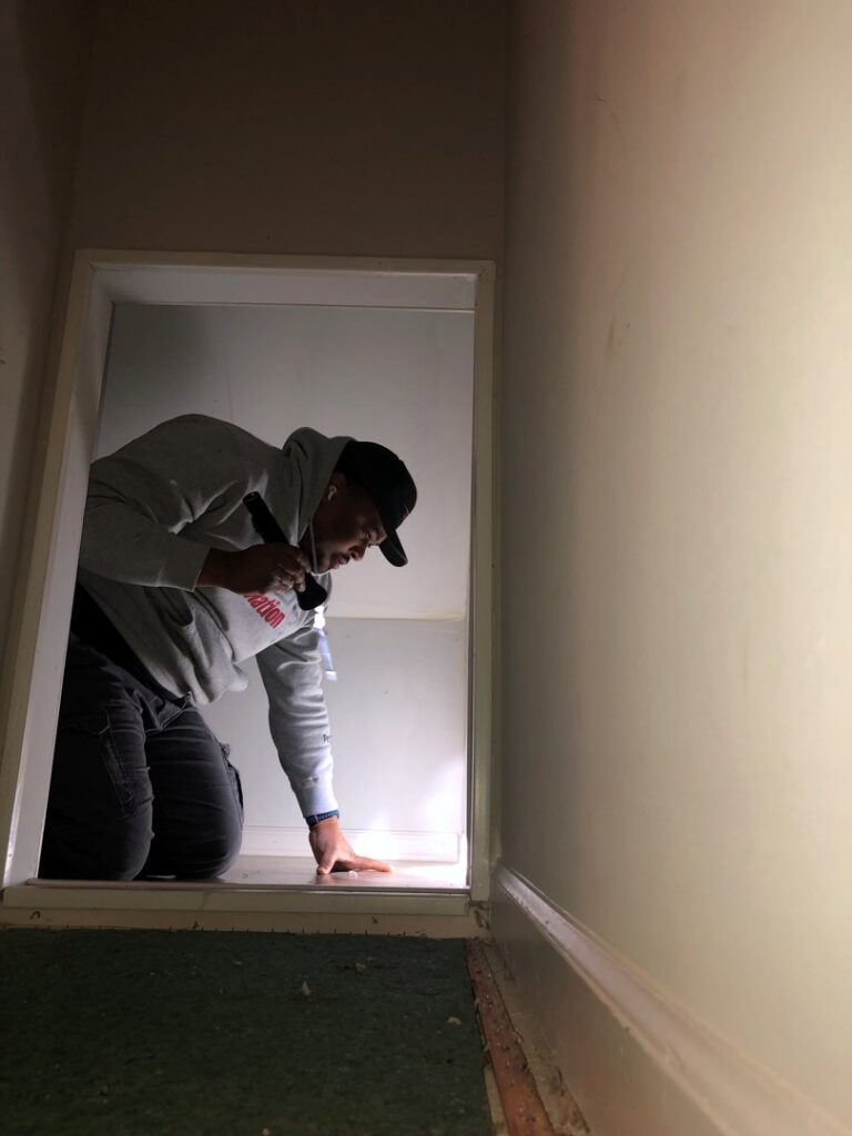 A PestNation technician inspecting a small access point with a flashlight for pest entry in Buford, GA