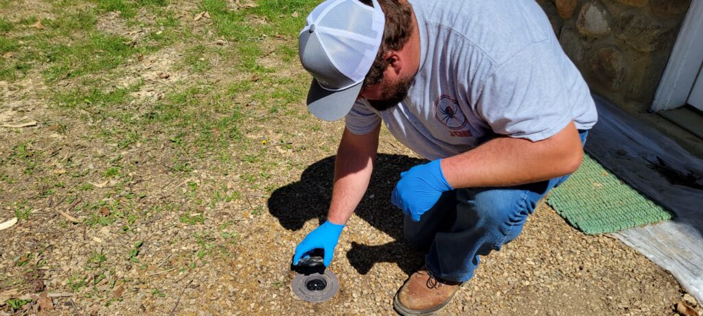 A pest control technician inspecting a ground bait station for rodents or insects, part of a service by Pest In Class Exterminating in Johnson City, TN.