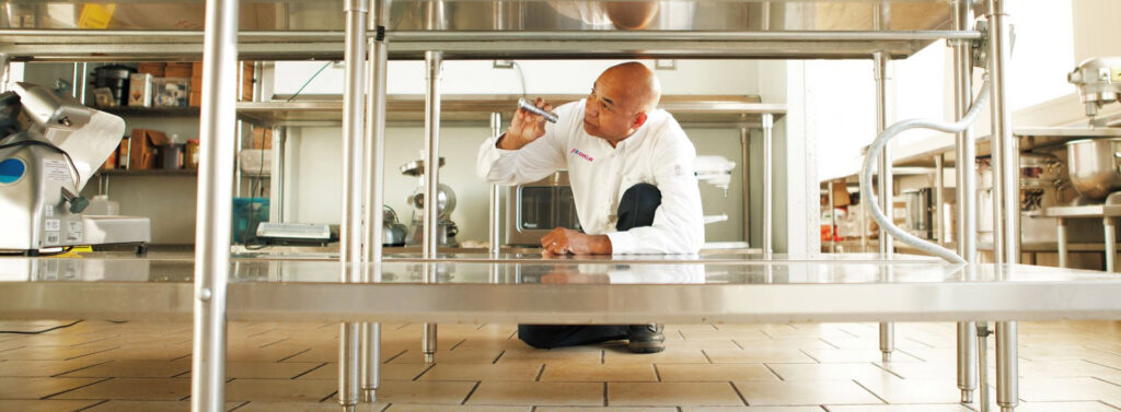 A Franklin Pest Solutions technician inspecting under a stainless steel table in a commercial kitchen in Lafayette, IN.