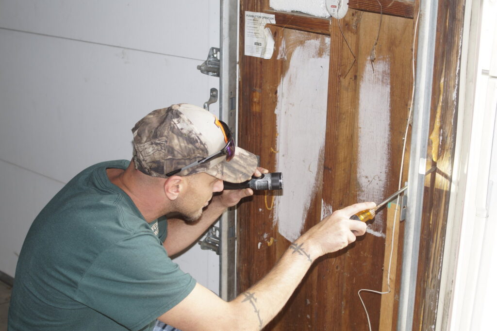 A pest control technician inspecting a wooden garage door frame with a flashlight for signs of pests or damage by Midwest Pest Control in East Peoria, IL.