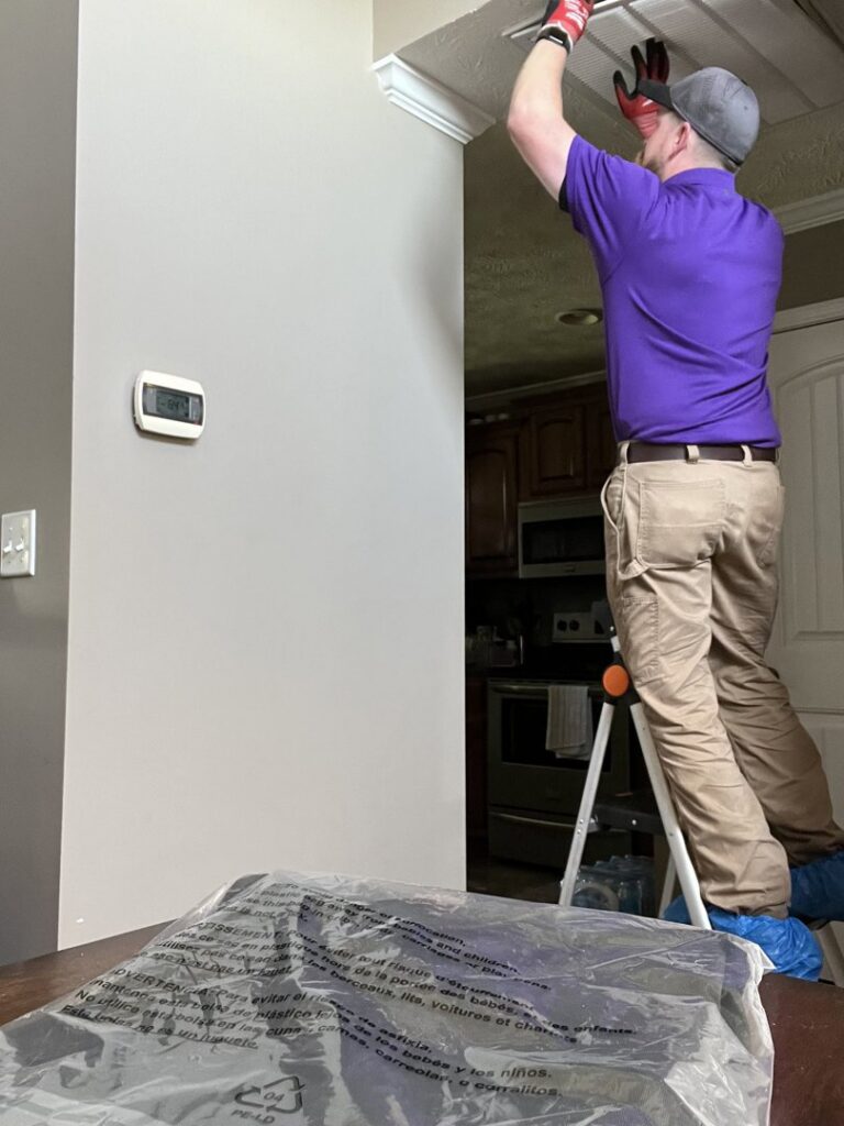 A Milan Heating & Air technician inspecting an indoor HVAC vent on a ladder in Milan, TN.