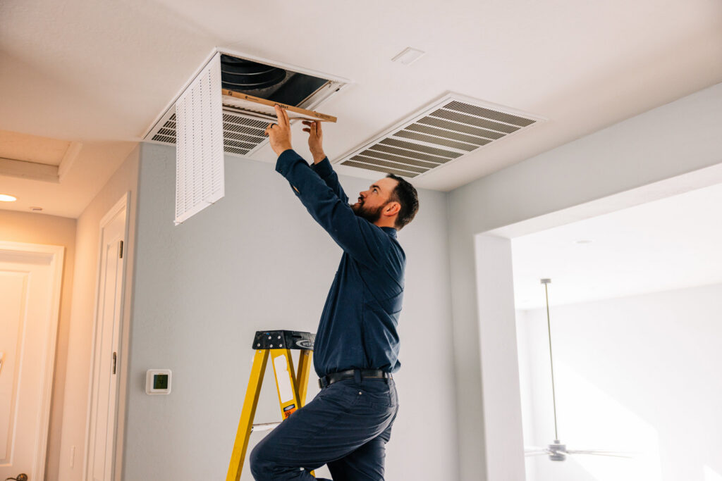 A technician from Rusty's Air Conditioning And Heating inspecting an indoor air vent or return on a ladder in Mesa, AZ.