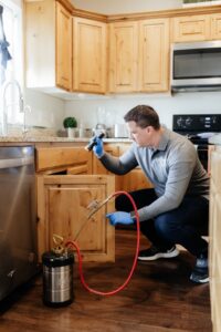 A pest control technician from BAM Pest Control in Sandy, UT, inspecting under a kitchen sink cabinet with a flashlight.
