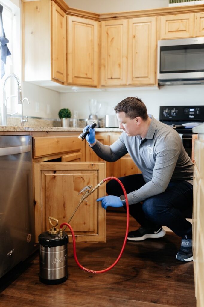 A pest control technician from BAM Pest Control in Sandy, UT, inspecting under a kitchen sink cabinet with a flashlight.