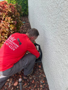 A Paske Pest Control & Wildlife Solutions technician inspecting a pest bait station near a building foundation in Cape Coral, FL.