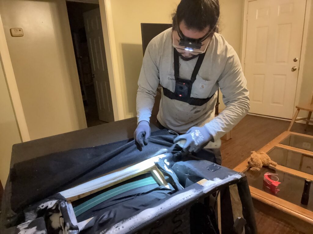 A pest control technician with a headlamp and gloves inspecting the underside of a sofa for pests by Tailor Made Pest and Wildlife in Louisville, KY.