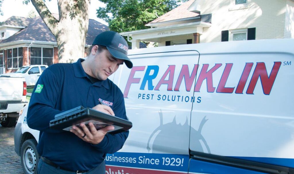 A Franklin Pest Solutions technician inspecting and writing on a clipboard next to a company van in Lafayette, IN.