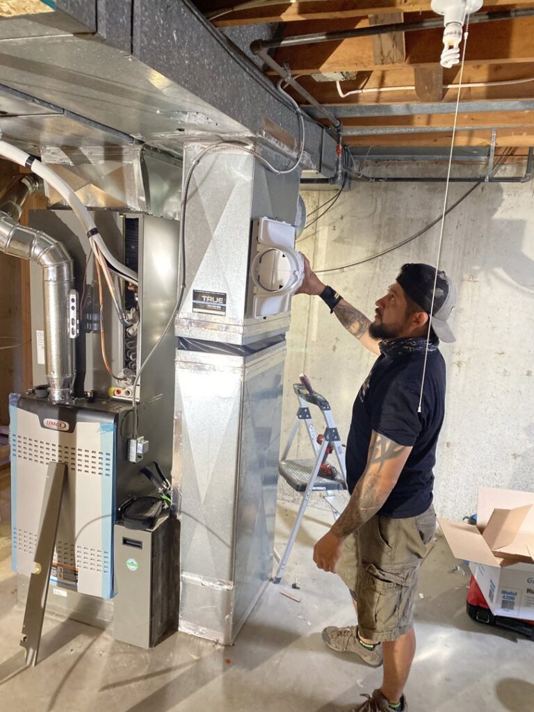 A technician installing an air purifier or humidifier on a furnace unit for True Heating and Cooling in Westminster, CO.