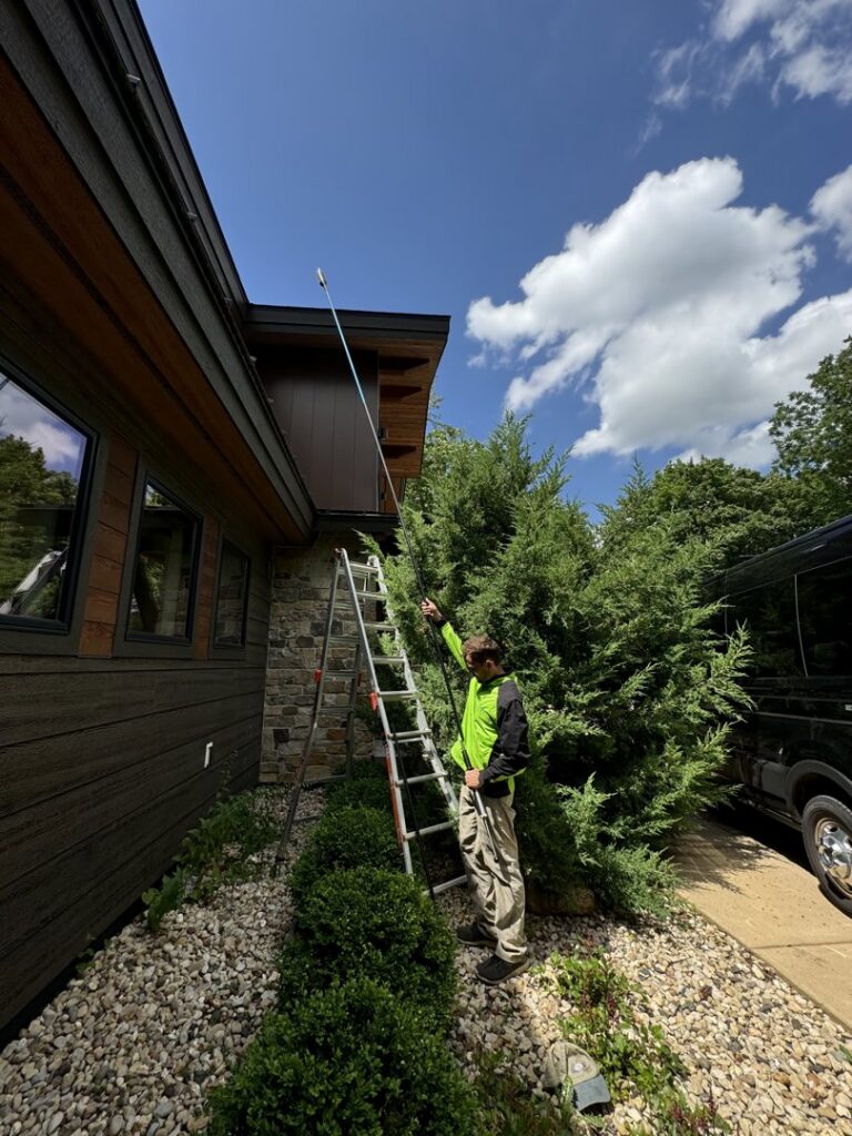 A Green Harmony technician on a ladder spraying the eaves of a house for pest control in Oconomowoc, WI.