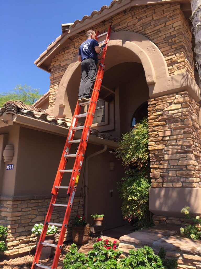 A technician on a tall ladder inspecting or working on the eaves of a residential building for Pigeon Control Phoenix AZ in Glendale, AZ.