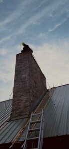 A Chimney Wizards LLC technician on a ladder, preparing to sweep a tall brick chimney on a roof in Orofino, ID.