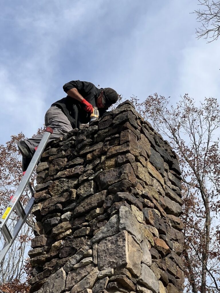 A technician on a ladder performing work on top of a stone chimney for Northwest Chimney in Bremerton, WA.