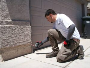 A pest control technician placing bait or inspecting a crack near a garage door for Advantage Pest & Weed Control in Rio Rancho, NM