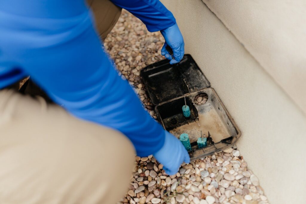 A pest control technician placing bait into an outdoor pest control station on a gravel bed for BAM Pest Control in Sandy, UT.