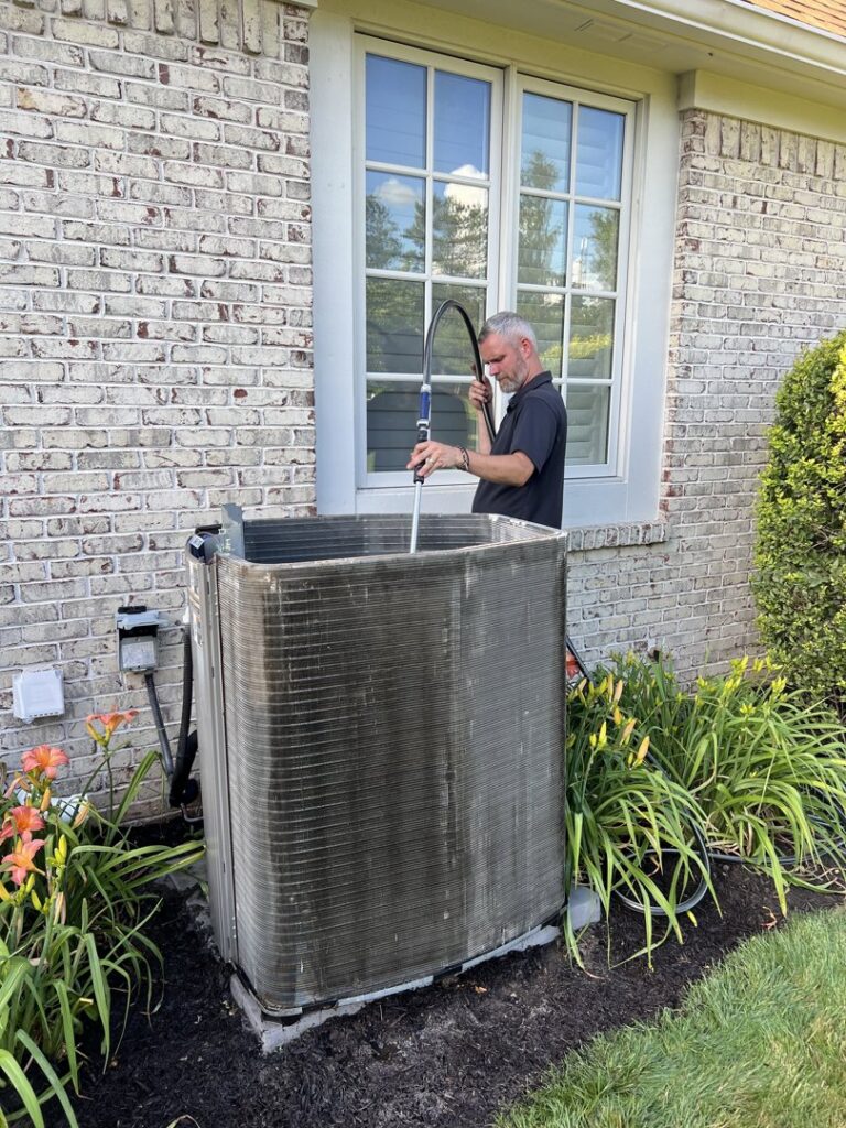 A technician power washing an outdoor AC condenser unit as part of routine maintenance by Radiant Heating & Cooling in Fishers, IN.