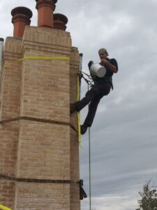 A technician rappelling down a tall brick chimney, performing maintenance or inspection for HBR Hearth and Chimney in Dallas, TX