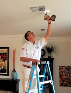 A Duct Doctor of Baton Rouge, LA technician on a ladder, carefully removing a ceiling air vent for cleaning or maintenance.