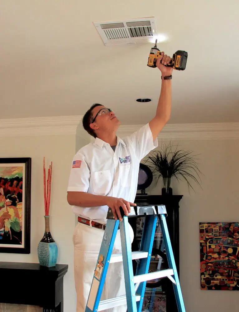 A Duct Doctor of Baton Rouge, LA technician on a ladder, carefully removing a ceiling air vent for cleaning or maintenance.