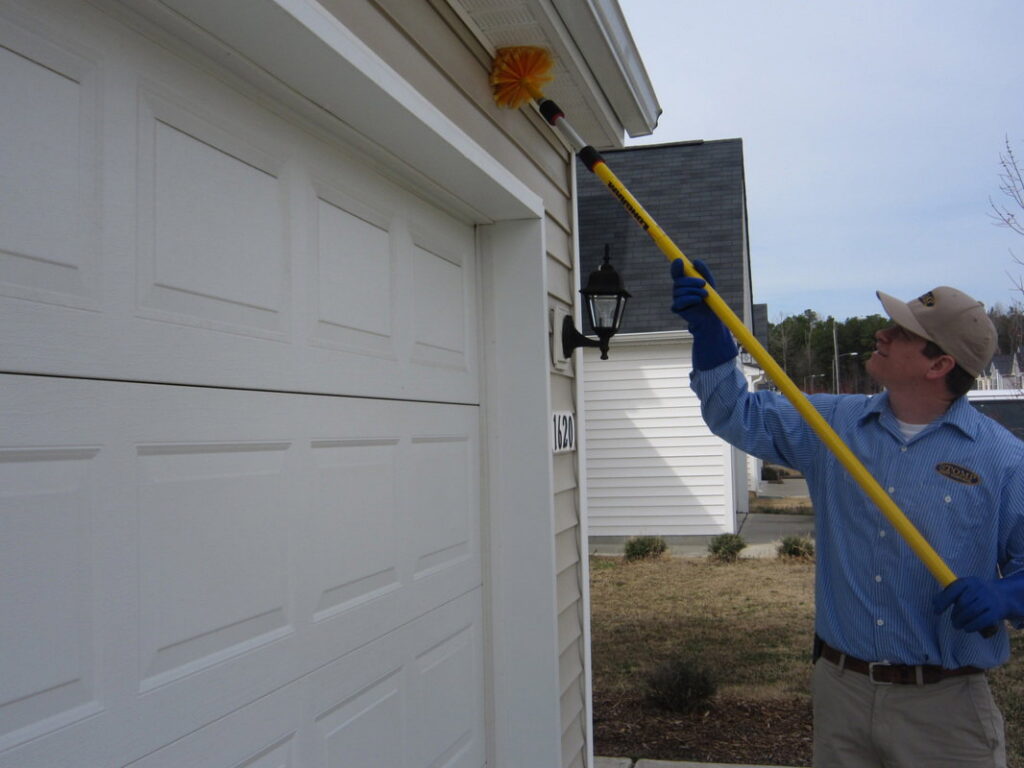 A Stomp Pest Control technician using a long brush to remove cobwebs from the eaves of a garage in Raleigh, NC.