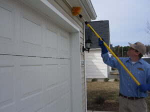 A Stomp Pest Control technician using a long brush to remove cobwebs from the eaves of a garage in Raleigh, NC.