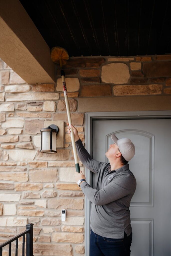 A pest control technician removing cobwebs or nests from the eaves of a house with a long-handled brush for BAM Pest Control in Sandy, UT.