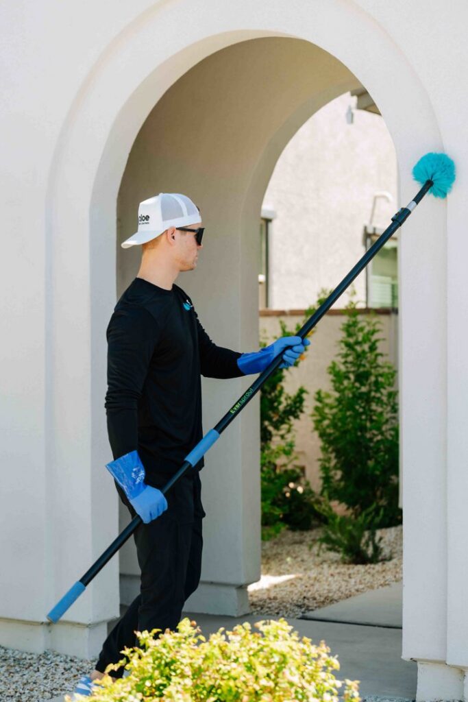 A pest control technician from Aloe Pest Control removing spider webs from a building exterior in Mesa, AZ.