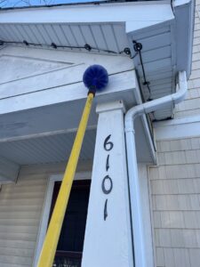 A technician using a long pole with a brush to remove spider webs from house eaves by Pike Creek Exterminating in Kenosha, WI.