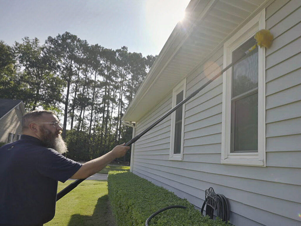 A Pest Magic Pest Control technician removing spiderwebs from house eaves with a long pole in Forsyth, GA.