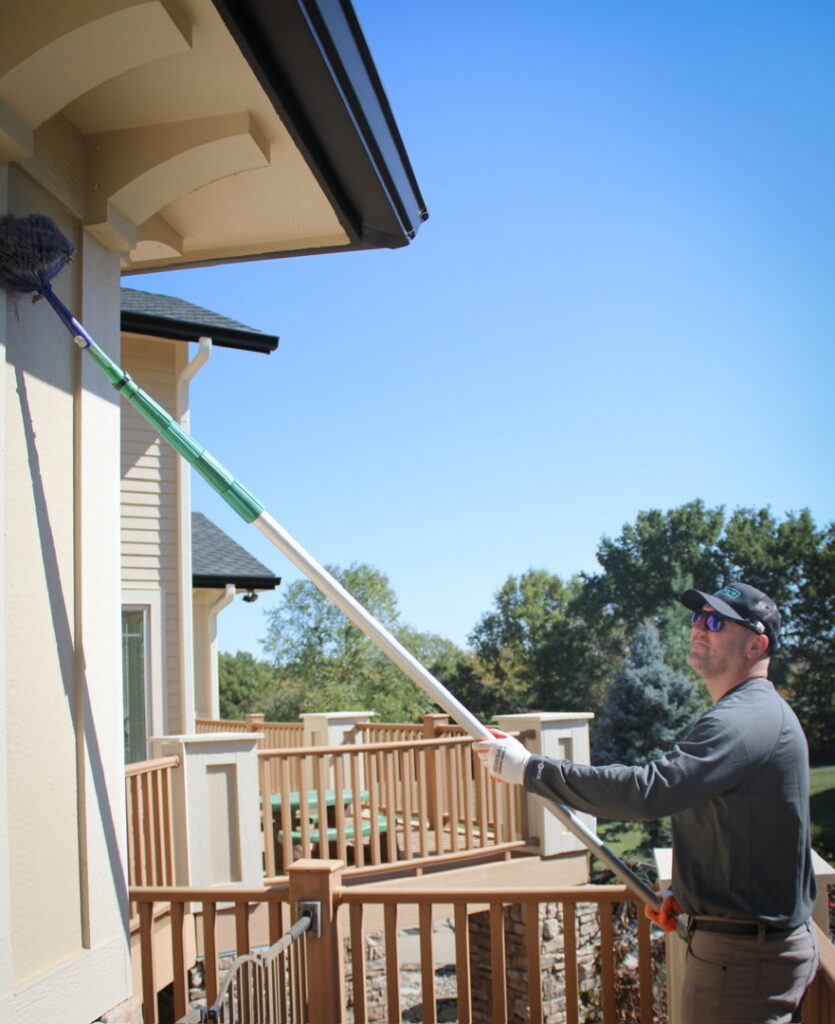 An Upfront Pest technician removing spiderwebs from a house corner above a deck in North Kansas City, MO.