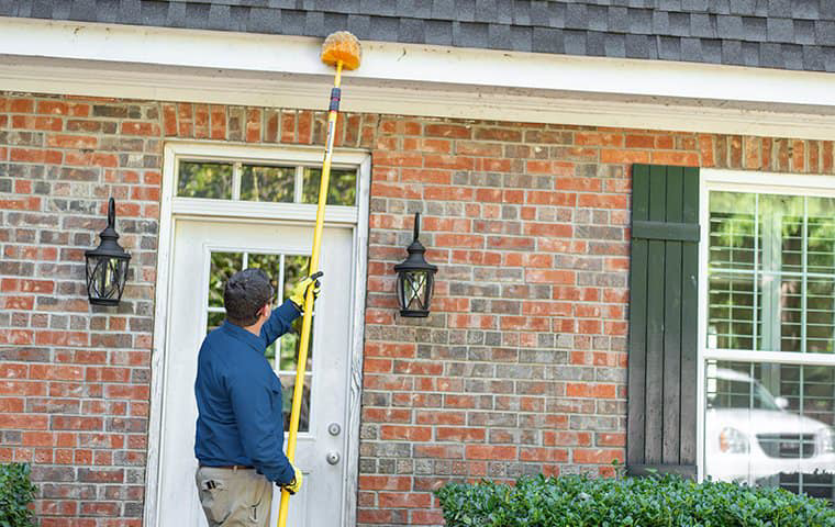 A pest control technician using a long pole to remove webs or nests from the eaves of a brick house for Pest Control Consultants in Sycamore, IL.