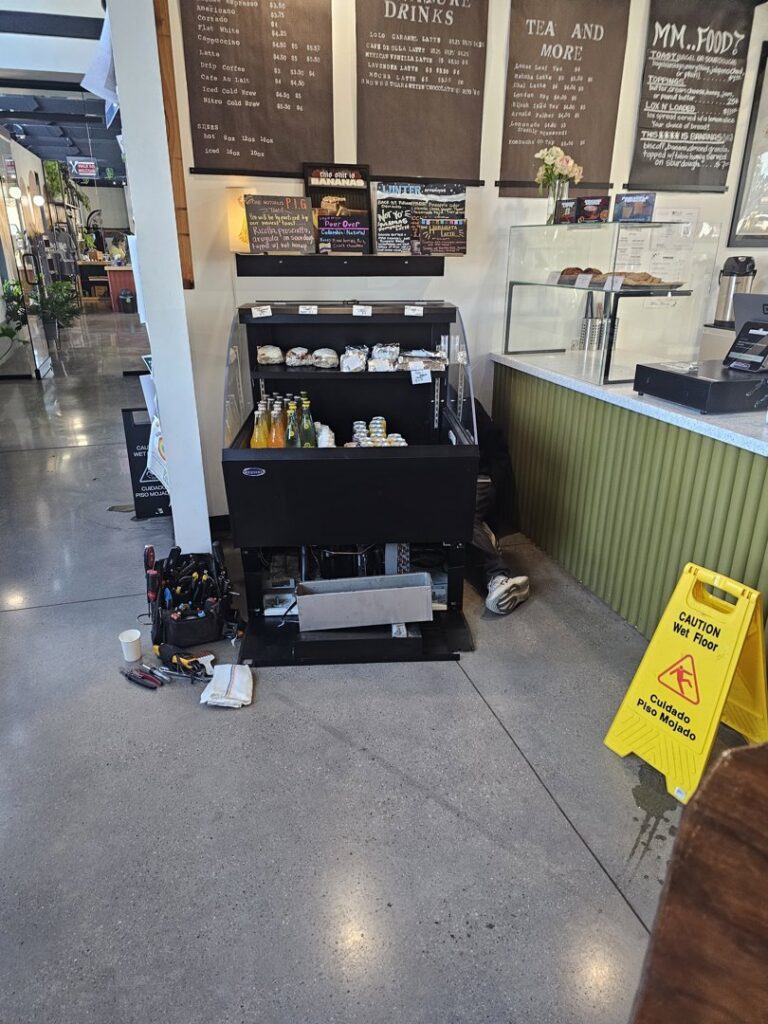 A technician repairing a commercial display refrigerator in a cafe, with tools laid out, by Henry Refrigeration & Hvac Utah in Ogden, UT.