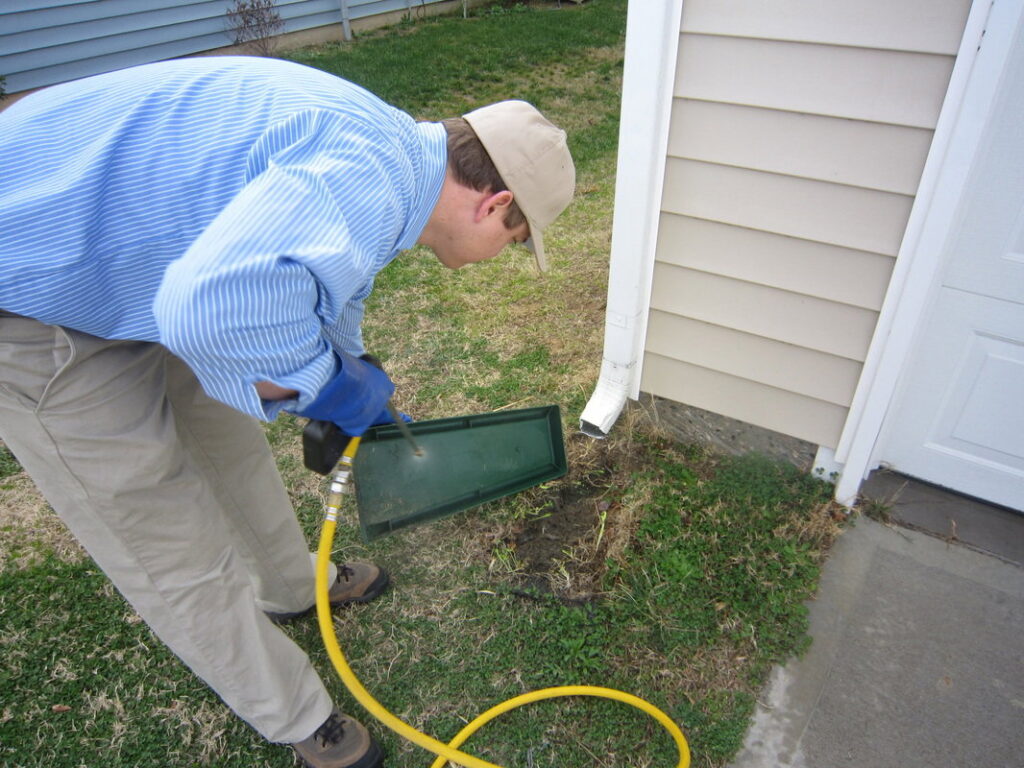 A Stomp Pest Control technician servicing a pest bait station near a residential foundation in Raleigh, NC.
