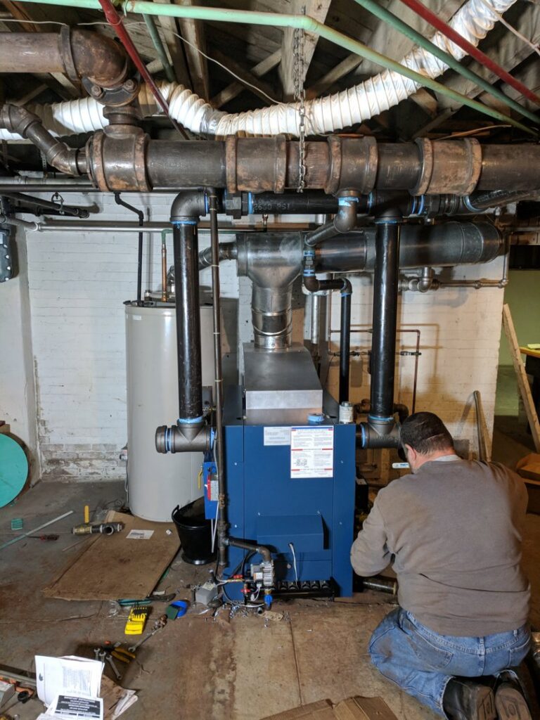 A Huether Heating & Cooling Inc technician servicing a large blue boiler system in a Rochester, NY basement.