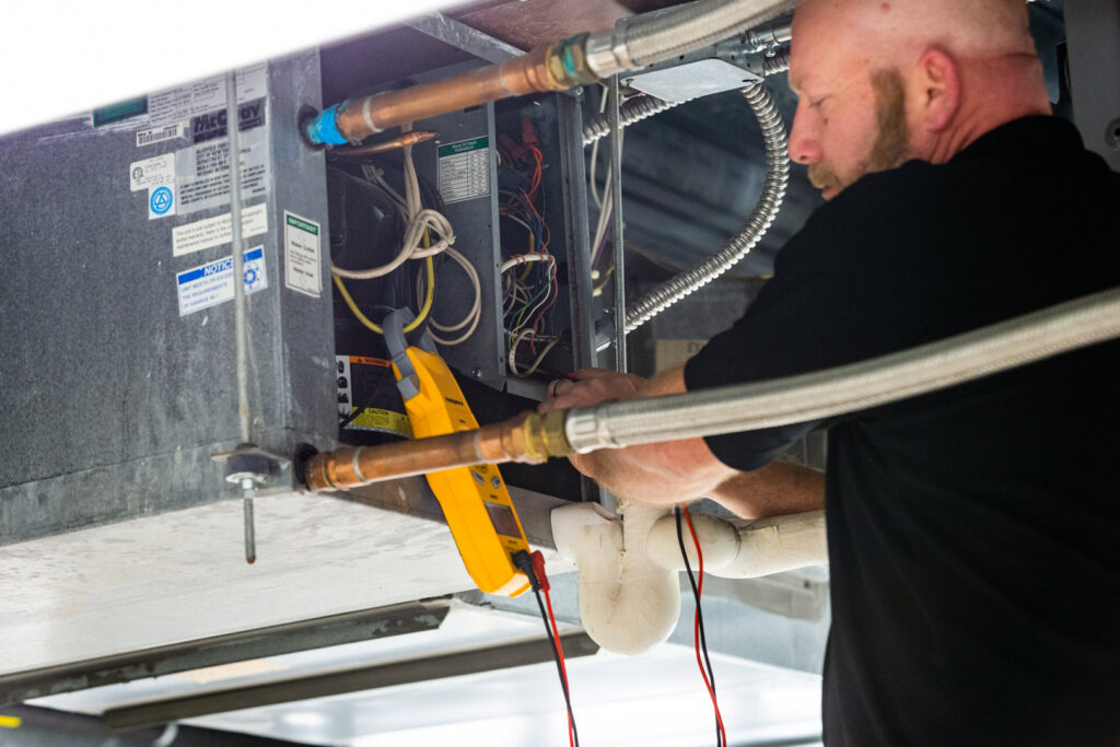 A KCM Heating & Air Conditioning technician servicing an indoor HVAC unit with a multimeter in Omaha, NE.