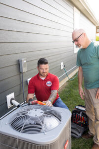 An S&B heating and cooling technician servicing an outdoor AC unit while a customer observes in Florence, KY.