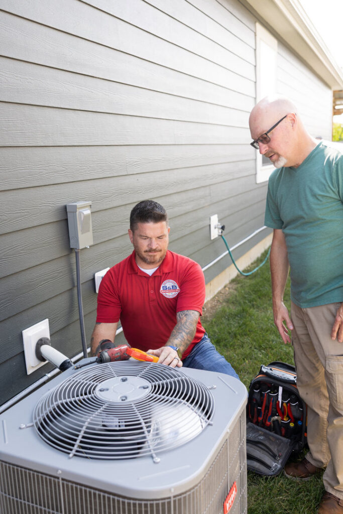 An S&B heating and cooling technician servicing an outdoor AC unit while a customer observes in Florence, KY.