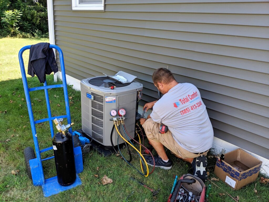 A technician from Total Comfort Heat and Air Conditioning, Inc. servicing an outdoor AC unit with gauges in Rochester, NY.