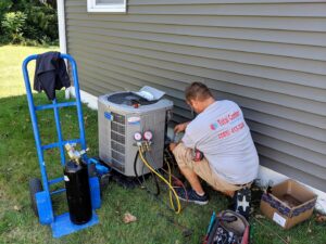 A technician from Total Comfort Heat and Air Conditioning, Inc. servicing an outdoor AC unit with gauges in Rochester, NY.
