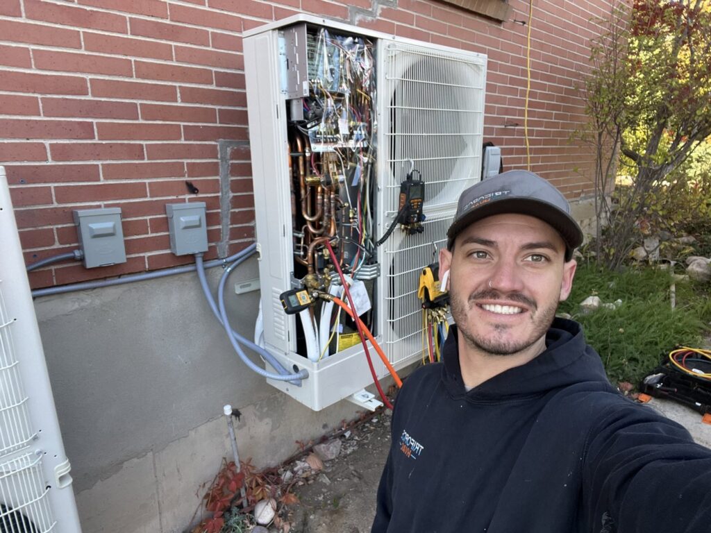 A TriForce HVAC technician smiling while servicing an outdoor HVAC unit, showing internal components and tools in Layton, UT