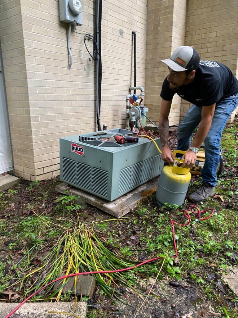 A technician servicing an outdoor RUUD air conditioning unit with gauges and a refrigerant tank for Adams Heating and Cooling in Schenectady, NY